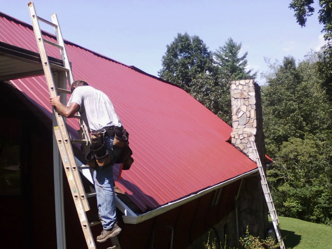Roofer on a ladder beside a red metal roof and stone chimney — Soffit & Fascia Repair by Roofing Brothers in Paden City